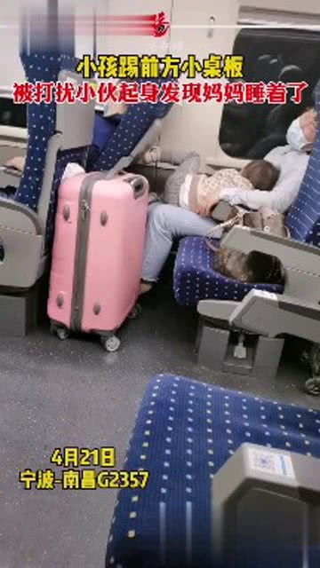 在一辆列车上,小孩用脚踢小桌板,前方男子被打扰后起身 